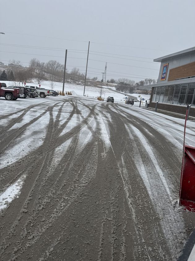 Snow-covered parking lot with tire tracks, parked cars, and a building on the right.