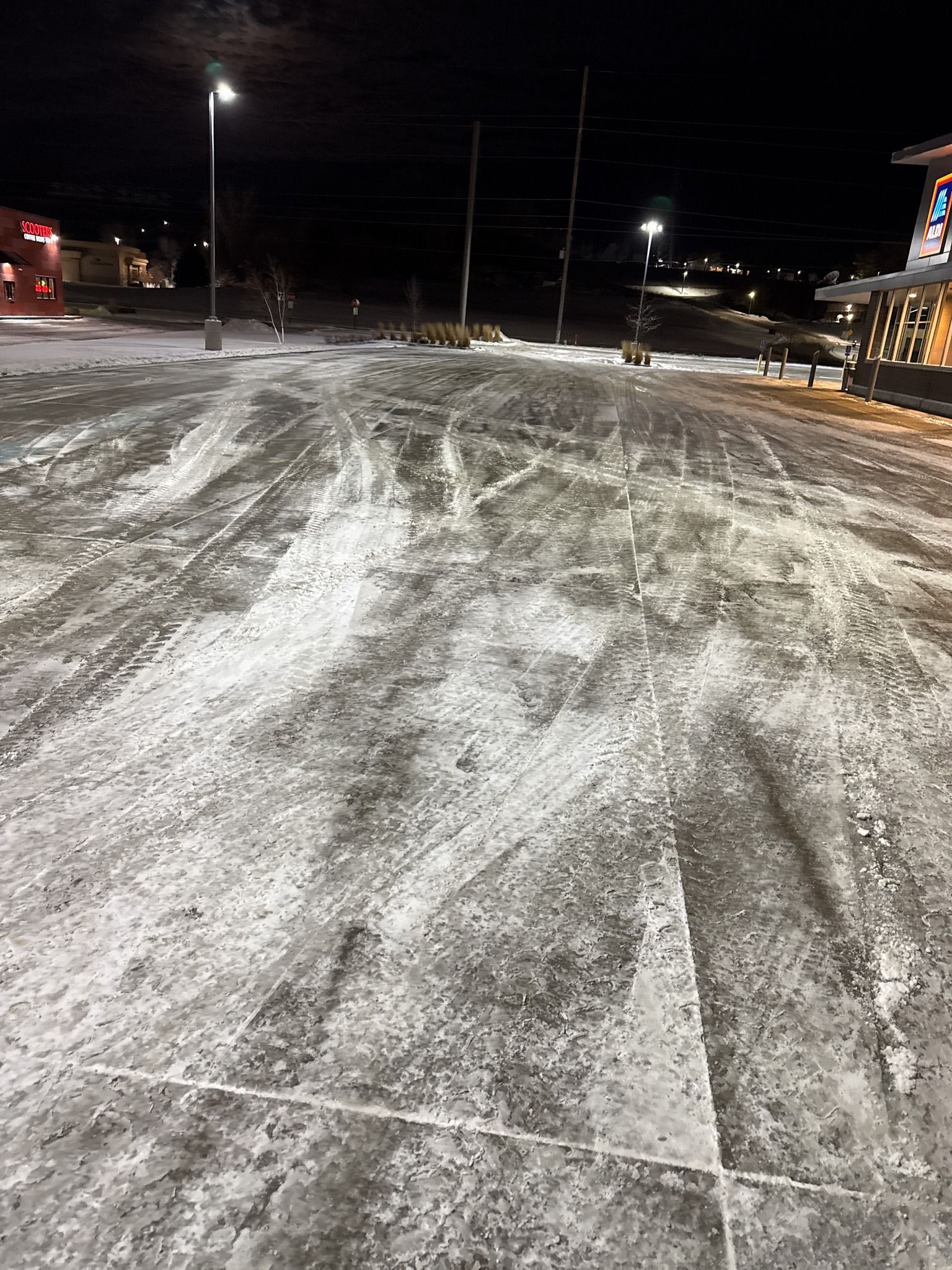 Snow-covered parking lot at night with tire tracks and lit storefronts in the distance