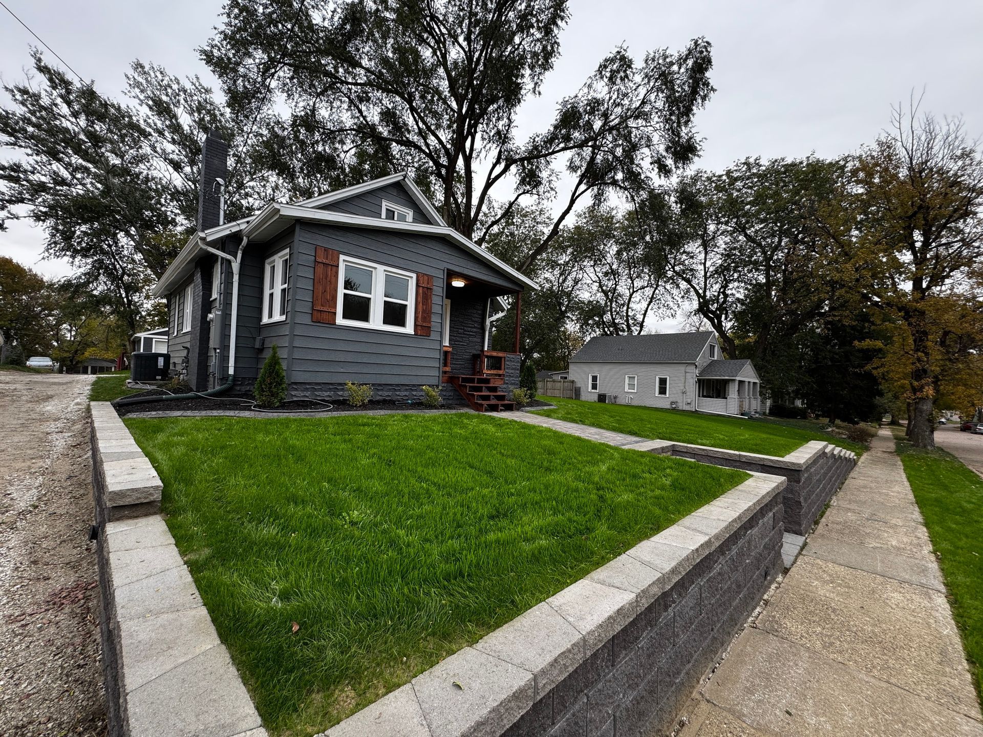Small gray house with white trim on a sloped green lawn beside a sidewalk and trees