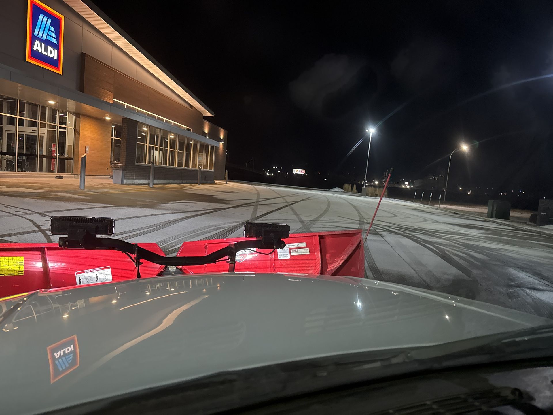 Snowplow scraping a snowy Aldi parking lot at night, viewed from inside a vehicle