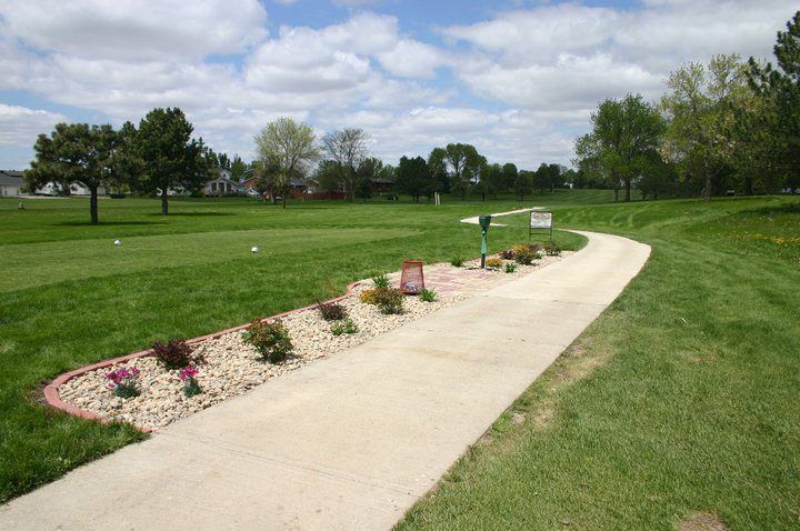 Curved concrete path through a grassy park with flower beds and a sign under a cloudy sky