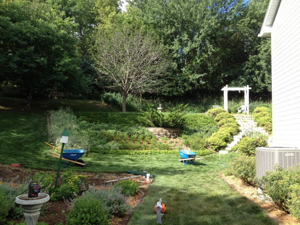 Backyard garden with green trees, shrubs, flower beds, and a small white pergola by the house
