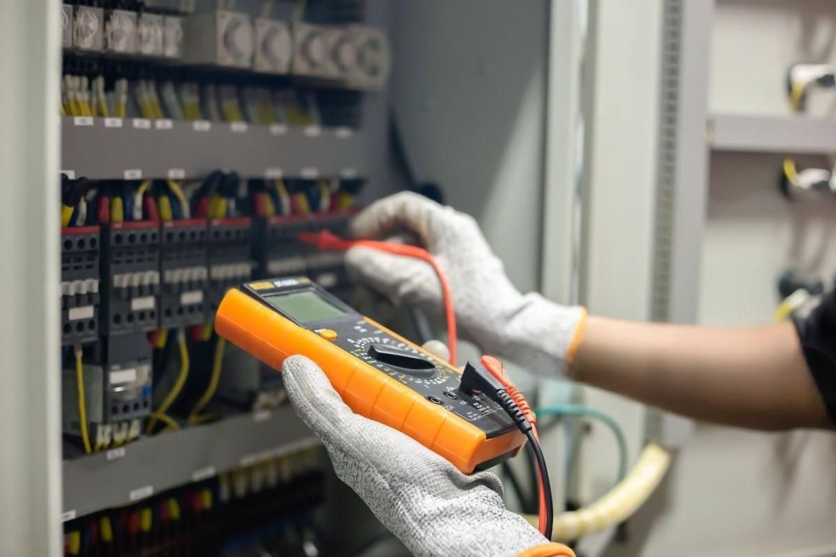 Person in gloves using a multimeter to test electrical wiring in a panel.