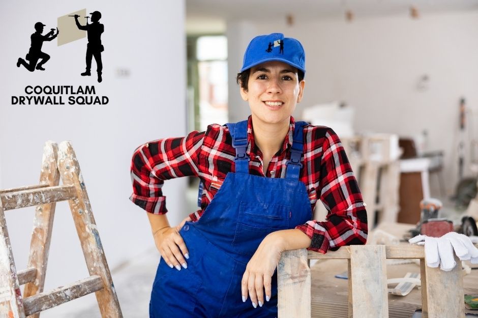 A professional drywall repair company worker wearing a blue cap, plaid shirt, and overalls poses confidently in a commercial renovation space. She leans casually against a wooden ladder with construction materials and tools in the background near Esplanade Ave, and Mission Rd in North Vancouver, BC.