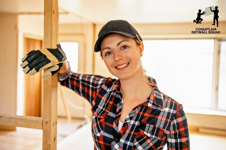 A drywall repair company worker smiles while leaning against wooden framing in a residential construction site. She wears a plaid flannel shirt and baseball cap, positioned in an unfinished room with exposed beams and bright natural lighting near 188 St, and 124a Ave, Pitt Meadows, BC.