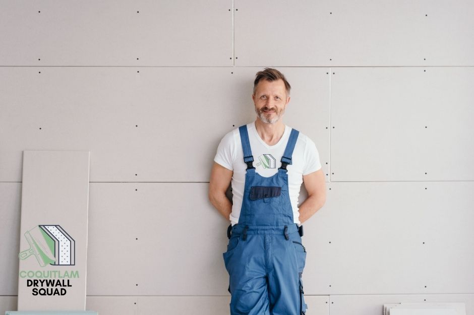 A drywall repair company professional man in blue overalls and a white t-shirt stands smiling against a wall with arms behind his back in a residential construction site, ready to provide expert drywall services near W 14th Ave, and Stephens St, Vancouver, BC.