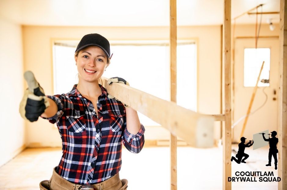 A drywall repair contractor in a plaid shirt and cap smiles while holding wood in an unfinished room with exposed wooden framing and drywall panels. She appears confident and ready to work on the residential construction project near Waresley St, and Thorne Ave in Maple Ridge, BC.