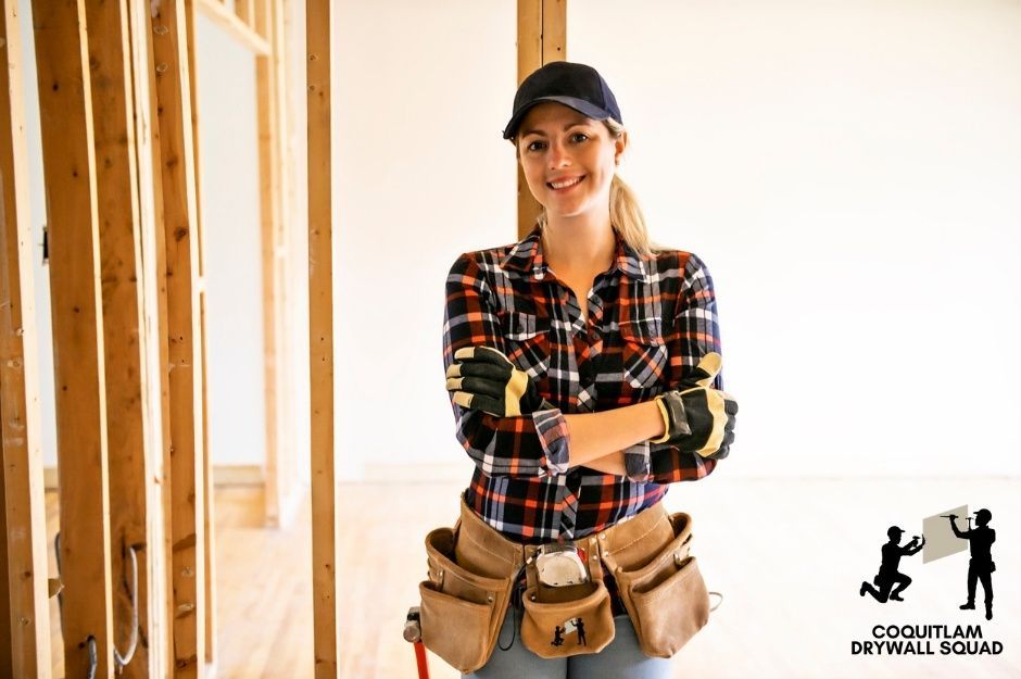 A professional drywall repair contractor stands confidently with arms crossed in a residential construction site. She wears a plaid flannel shirt, a work cap, and a tool belt around her waist. Behind her are exposed wooden wall studs of an unfinished room near 80 Ave, and 208 St in Willoughby, BC.