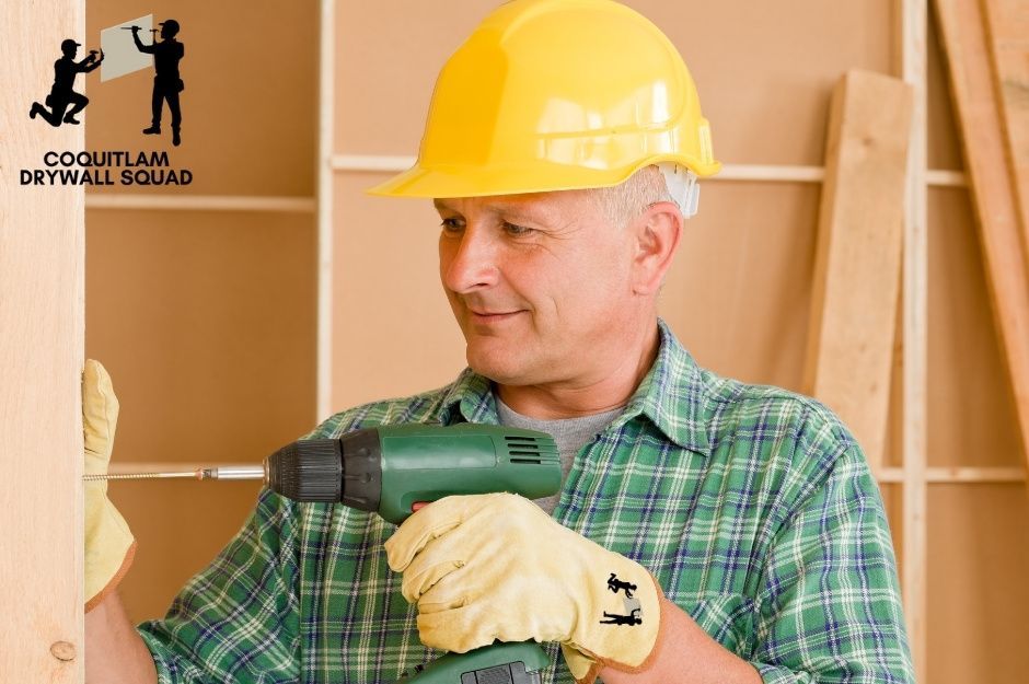 A professional drywall repair handyman wearing a yellow hard hat and green plaid shirt works on a residential project. He has on work gloves and is positioned among wooden framing near 54 St, and 5b Ave in Delta, BC.