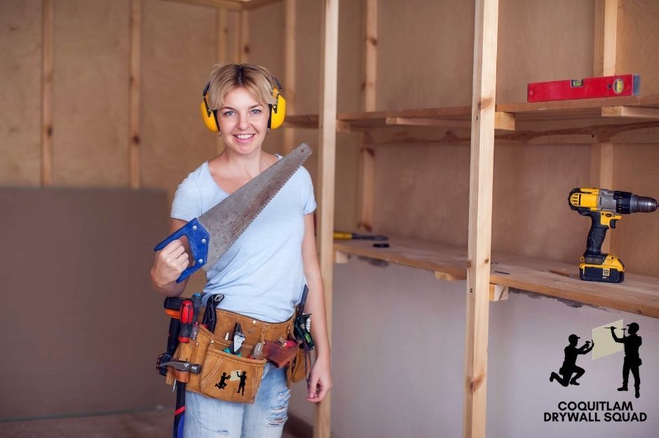 A drywall repair specialist wearing an ear protection and a tool belt stands in an unfinished room with exposed wooden framing. She holds a saw while wooden shelving is visible in the background near Fraser Hwy, and 206 St in Langley, BC.