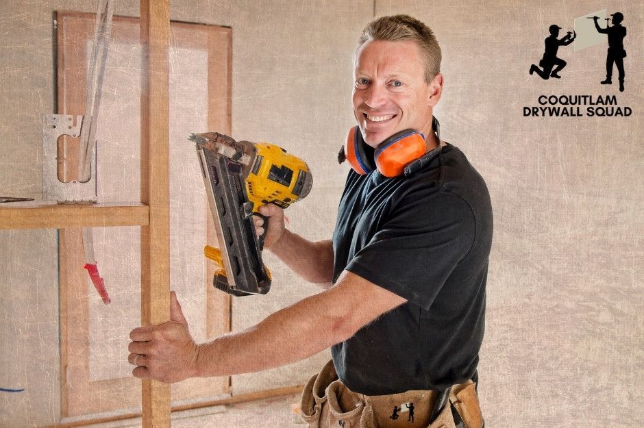 A professional drywall repair technician smiles while holding a yellow nail gun in a residential construction area. He wears a black shirt, tool belt, and safety gear, standing among exposed wooden studs and drywall sheets near Windermere St, and E 22nd Ave in South Vancouver, BC.