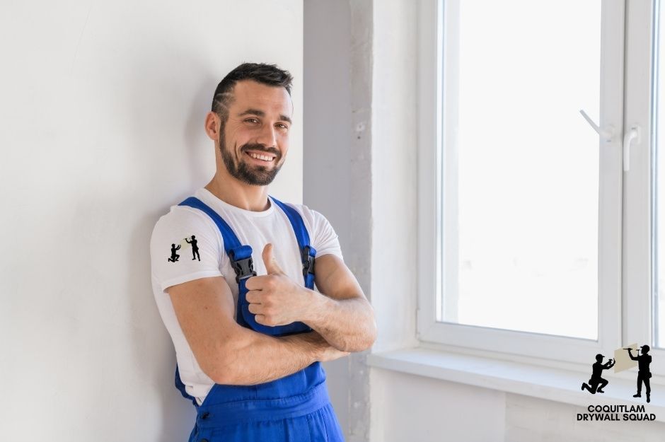 A drywall repair technician with a beard wearing blue overalls and a white t-shirt stands with arms crossed, smiling at a residential construction site with smooth white walls and a large window near 177b St, and 60 Ave, Surrey, BC.