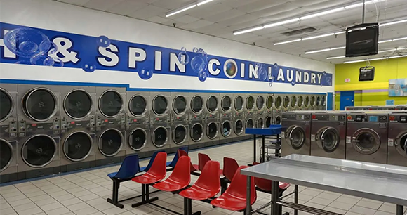 Inside a laundromat, rows of front-loading dryers line the wall beside red and blue chairs.