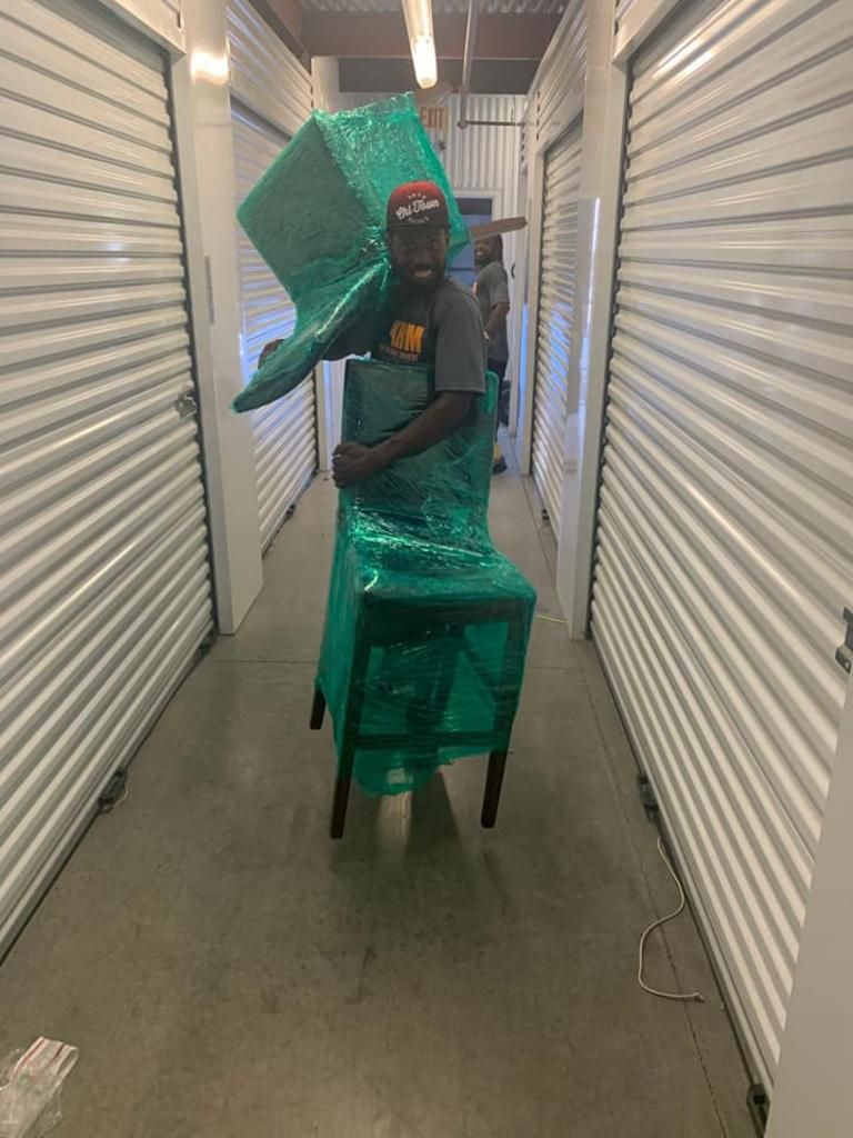 Person holding a chair wrapped in green plastic in a storage unit hallway.