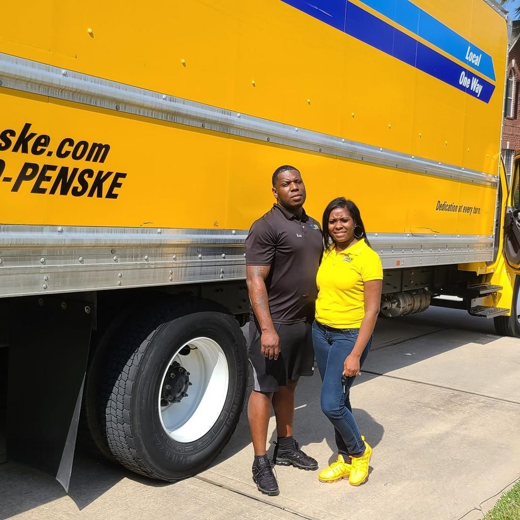 Two people pose in front of a yellow Penske truck.