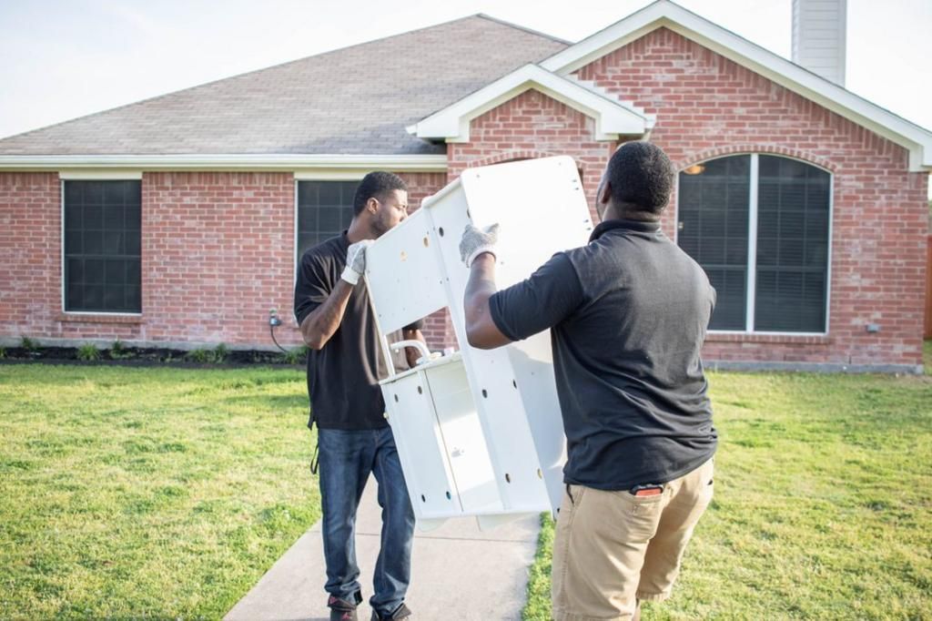 Two men carrying a white appliance towards a brick house on a sunny day.