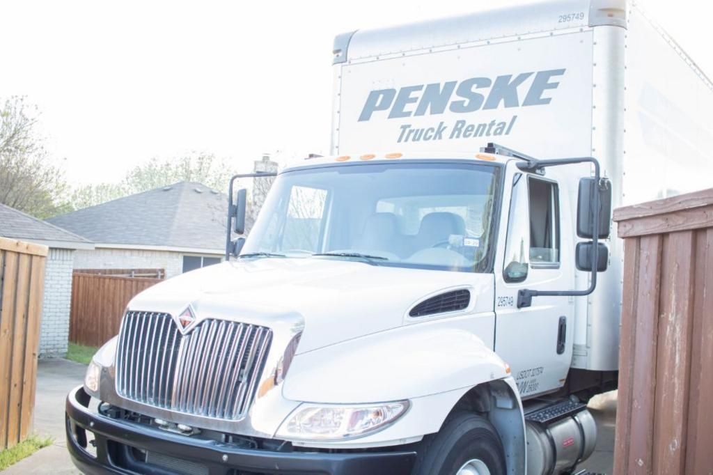 White Penske moving truck parked near a fence and small building.