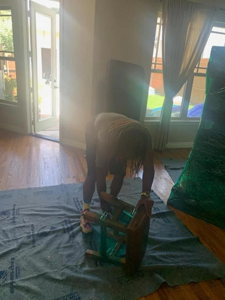 Person wrapping a chair in plastic, in a room with hardwood floors and large windows.