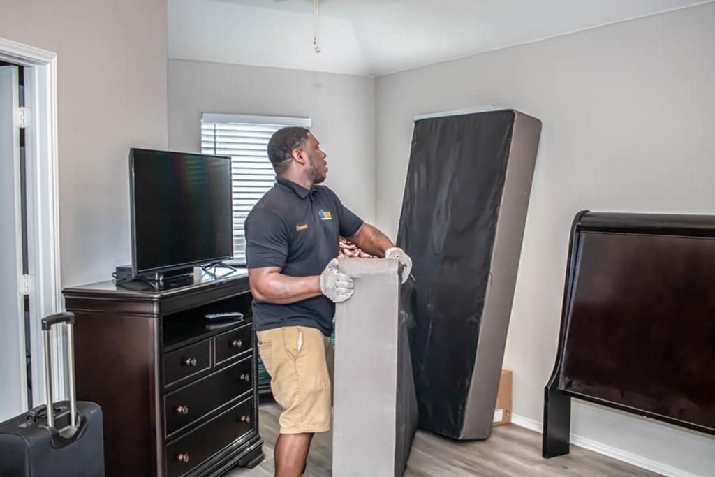 Man in gloves lifting a mattress in a bedroom, dresser with TV and luggage in view.