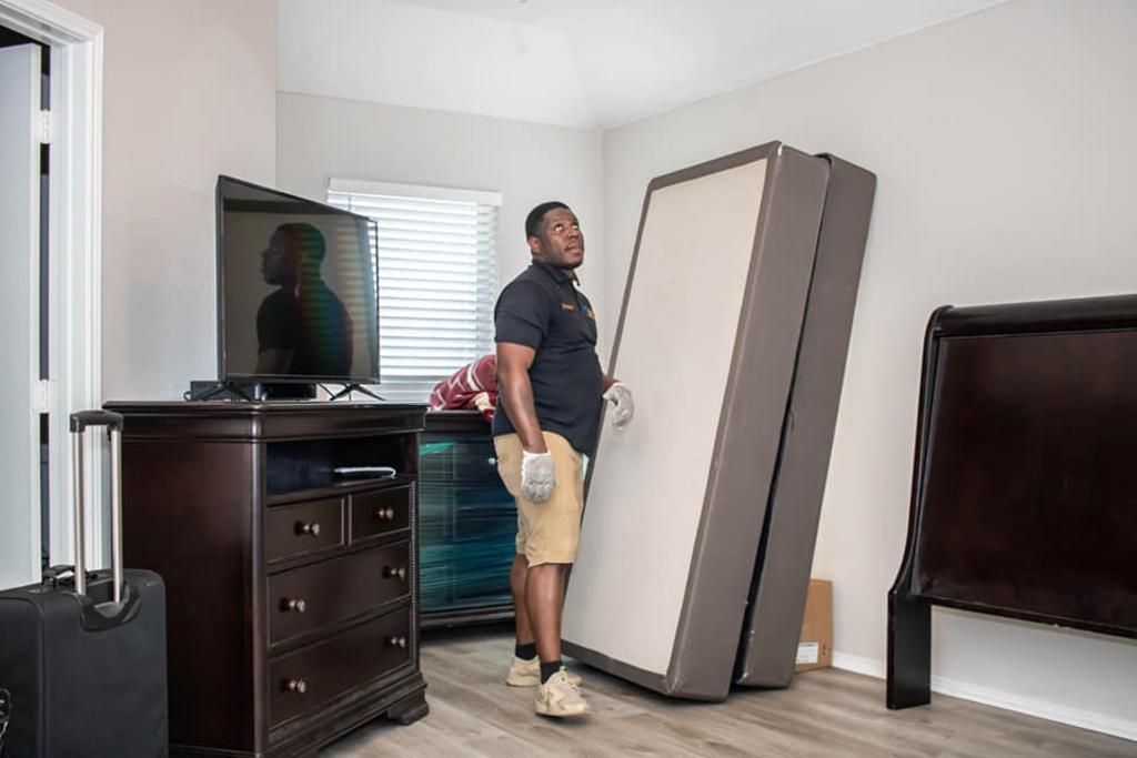 Man carrying mattress base in a bedroom, TV, dresser, and suitcase visible.