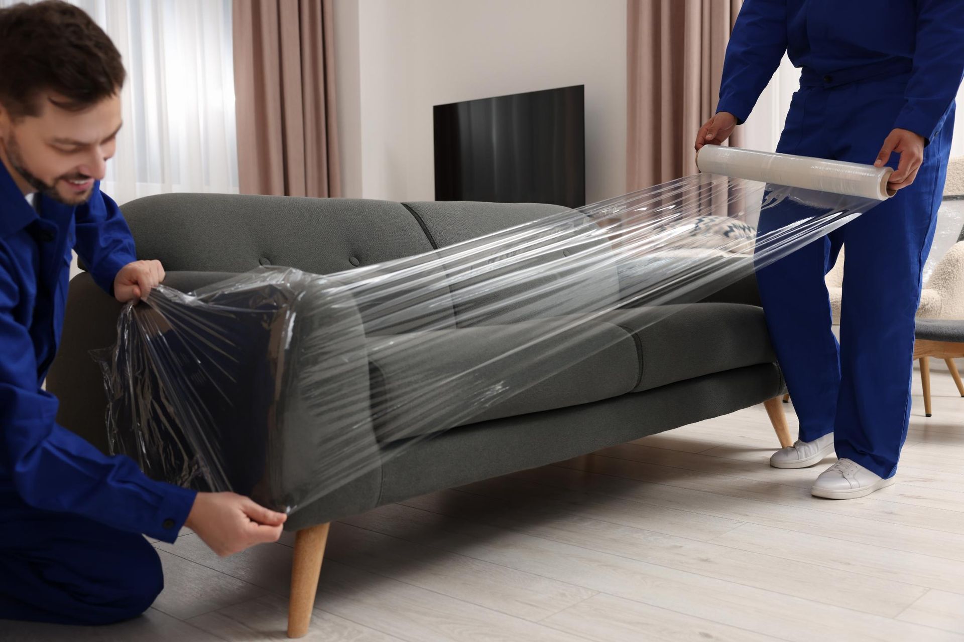 Two movers wrapping a gray couch in plastic wrap inside a living room.