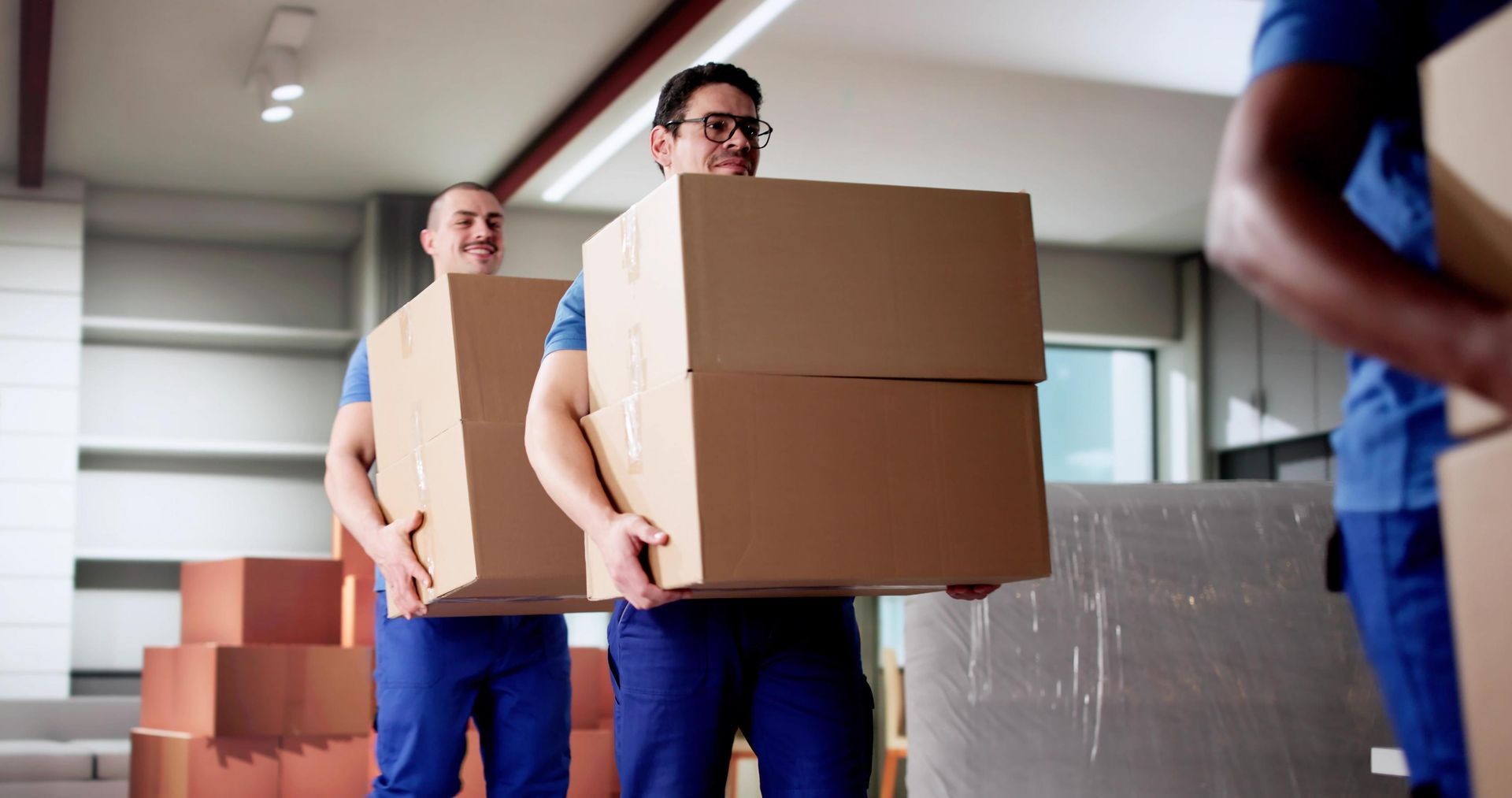 Movers in blue uniforms carrying stacked cardboard boxes inside a building.