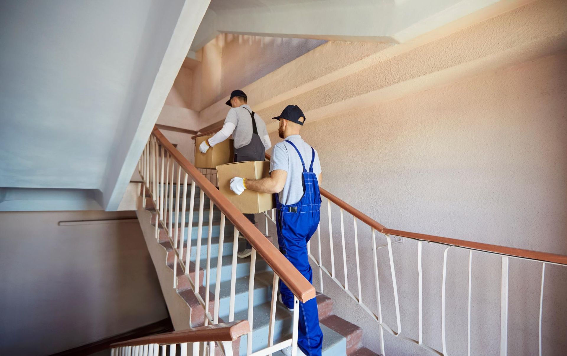 Two movers in blue coveralls carrying boxes up a stairwell.