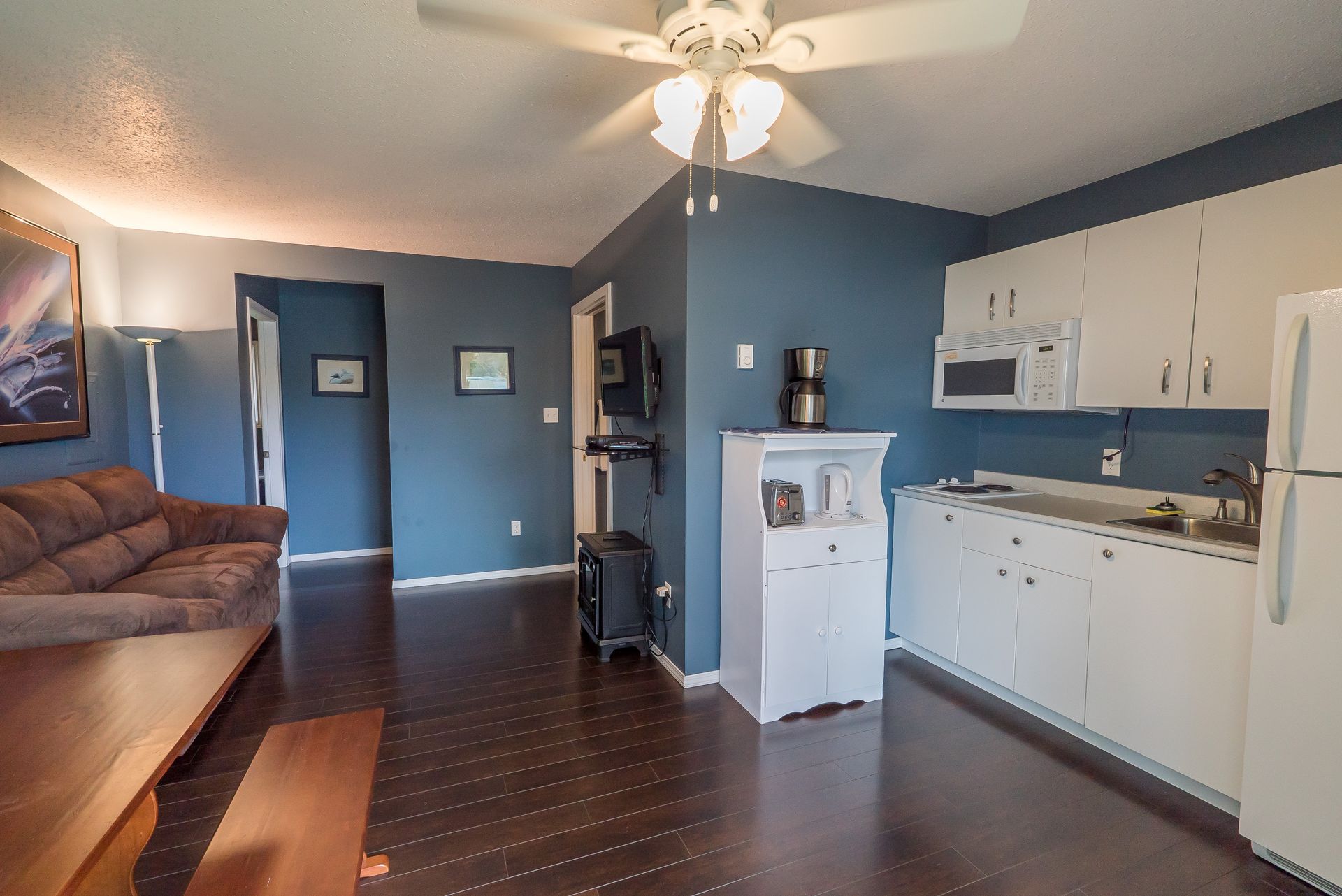 A room featuring a brown sofa, a wooden table, and a small kitchenette with white cabinets against dark blue walls.