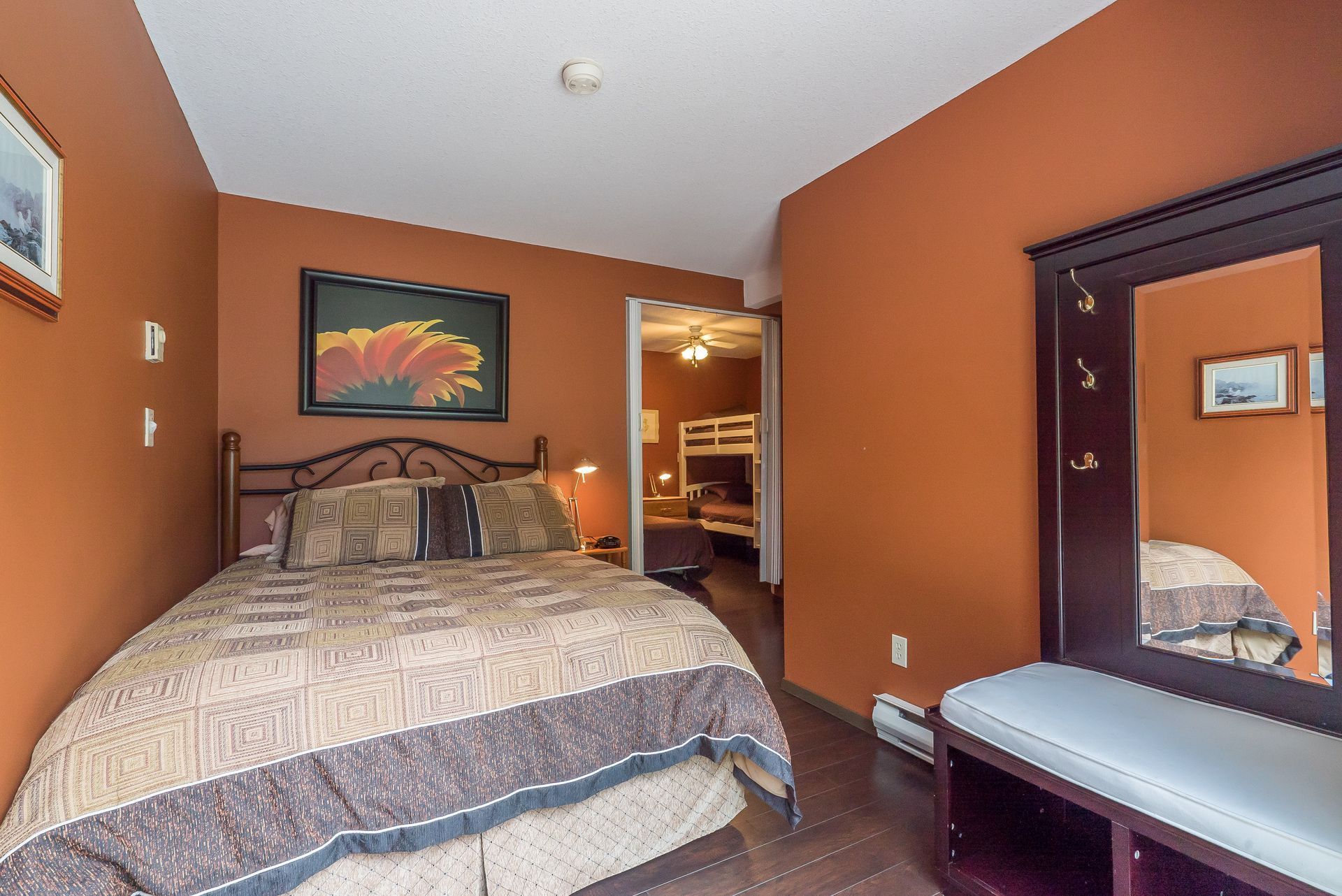 A hotel room with a queen bed, a painting of a flower, an entryway bench with hooks, and a view into a bunk bed room.