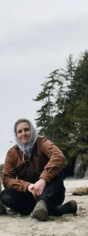 A person in a brown jacket and hooded sweatshirt sitting on a sandy beach with pine trees in the background.