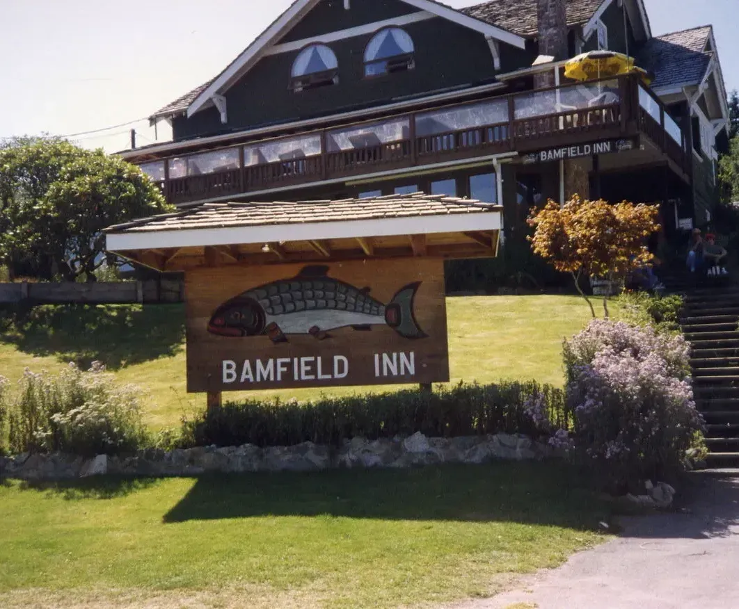 A wooden sign for the Bamfield Inn featuring a fish illustration, positioned in front of a green lodge on a grassy lawn.