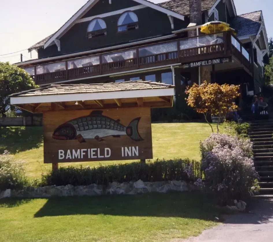 A wooden sign with a fish illustration in front of the Bamfield Inn, a dark green building with a large deck.