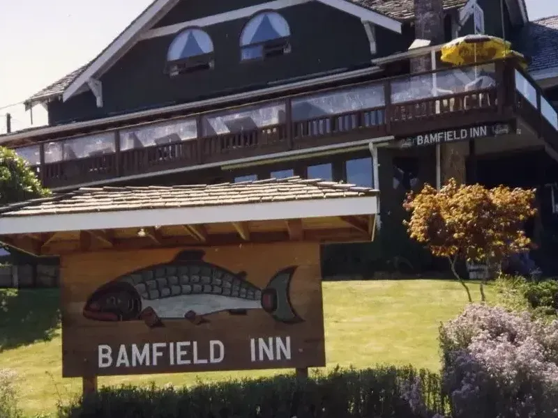 A wooden sign with a painted salmon graphic identifies the dark-sided Bamfield Inn, set against a green lawn and trees.