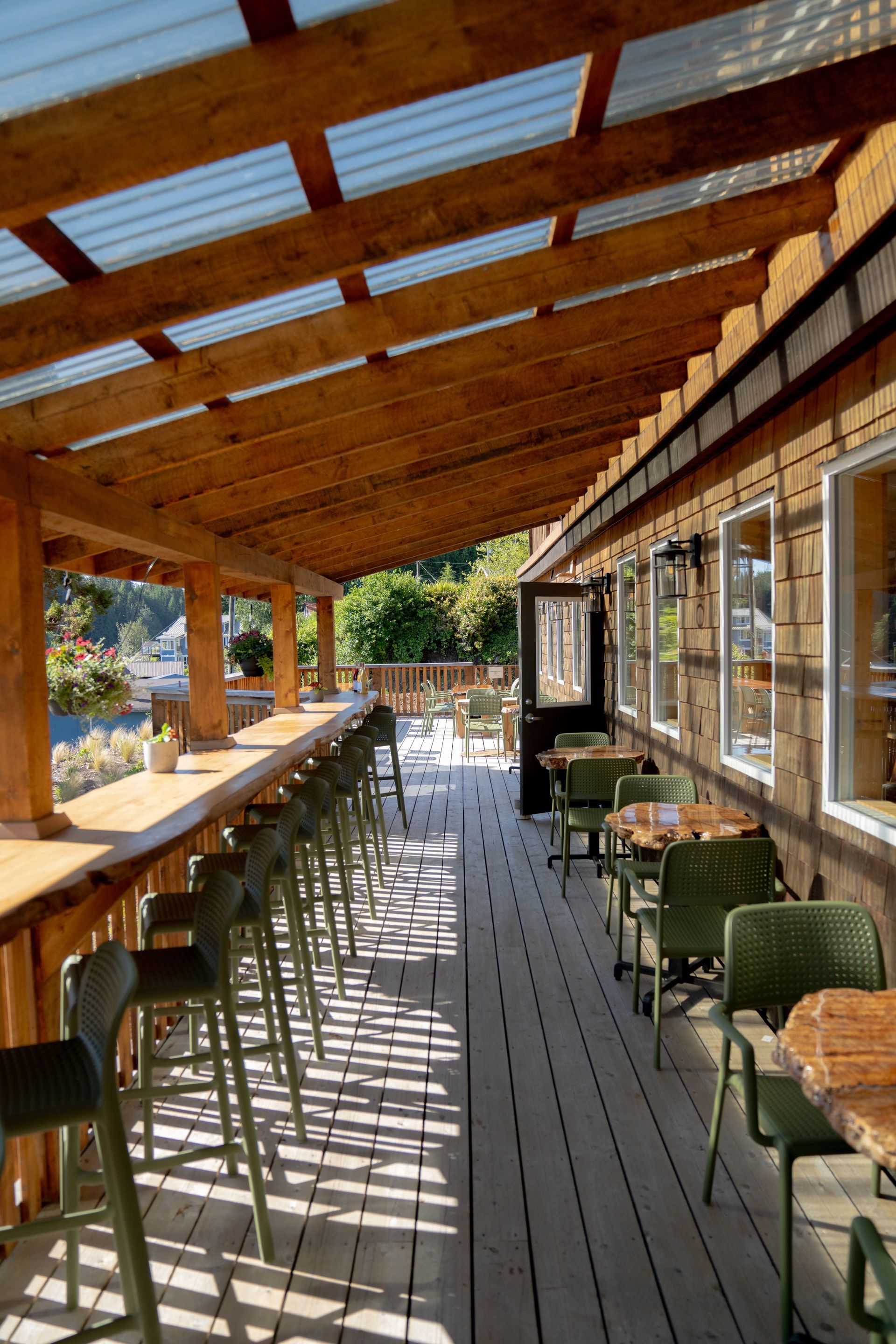 A rustic wooden deck features a long bar with stools and tables, sheltered by a translucent roof, overlooking greenery.