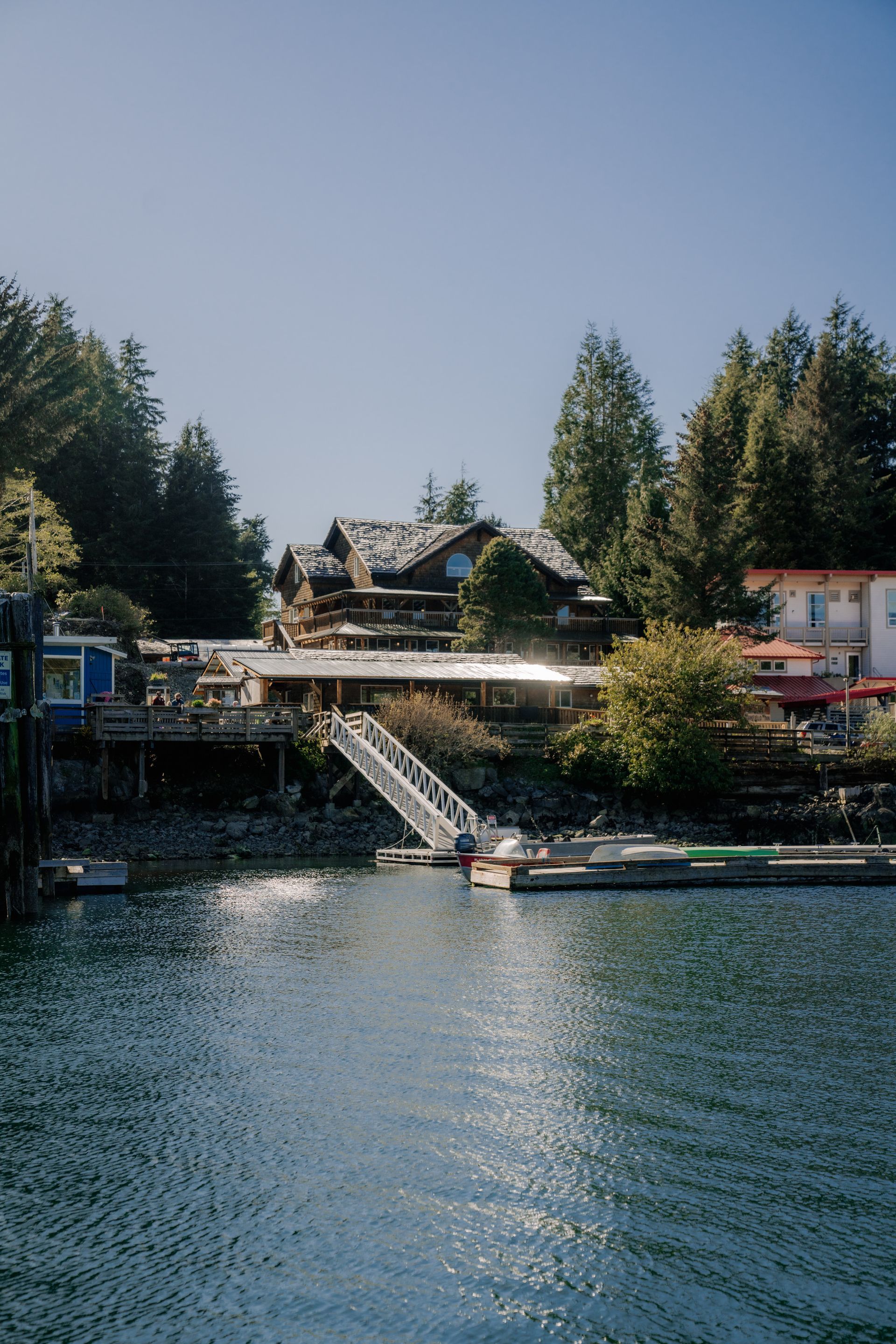 A wooden building sits on a hillside by a calm harbor, connected by a staircase to a floating dock.