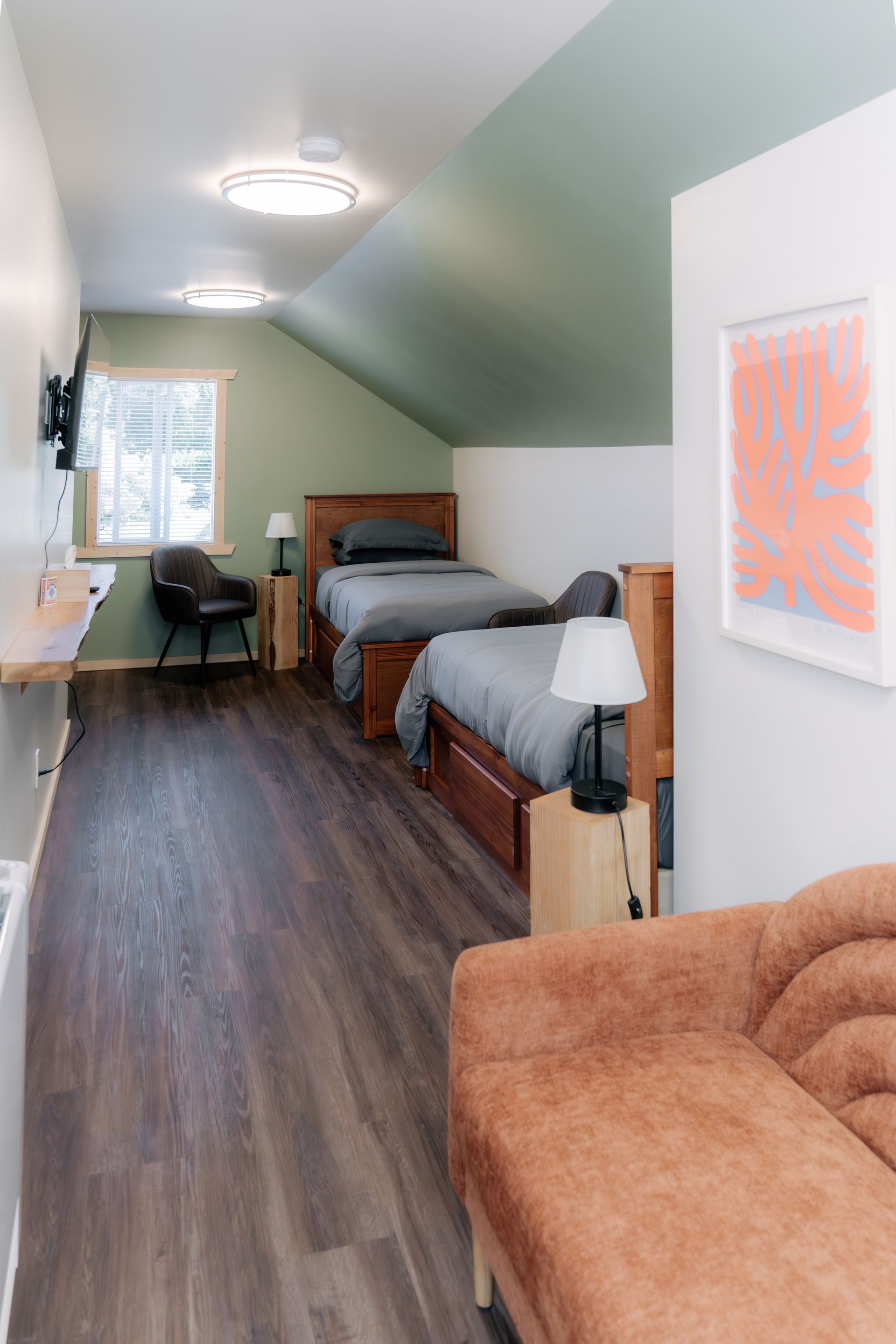 A room with two twin beds, dark wood flooring, sage green accent wall, a cozy sofa in the foreground, and a small desk.