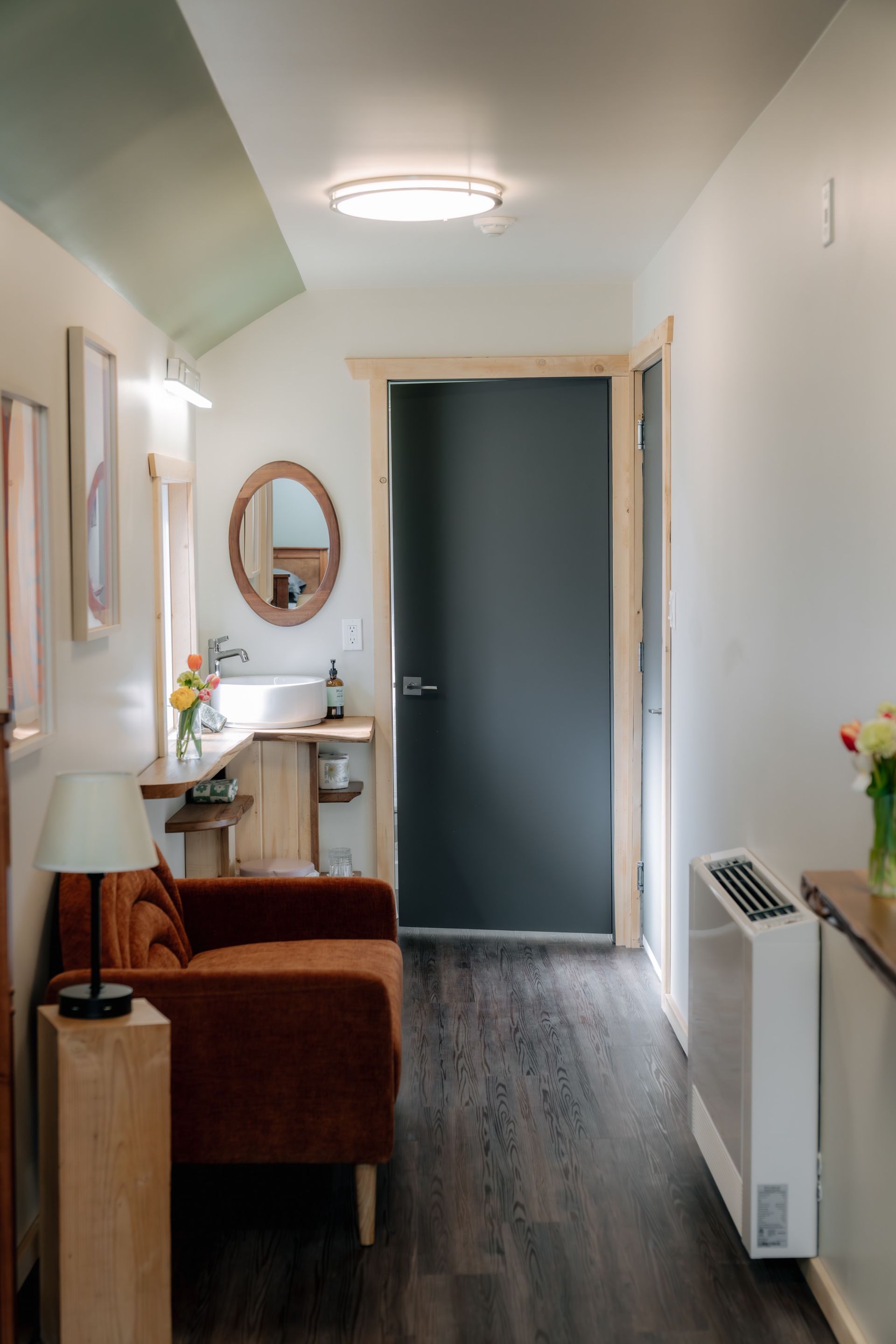 A narrow indoor hallway featuring a brown armchair, a lamp on a wooden stand, a small sink with a mirror, and a dark door.