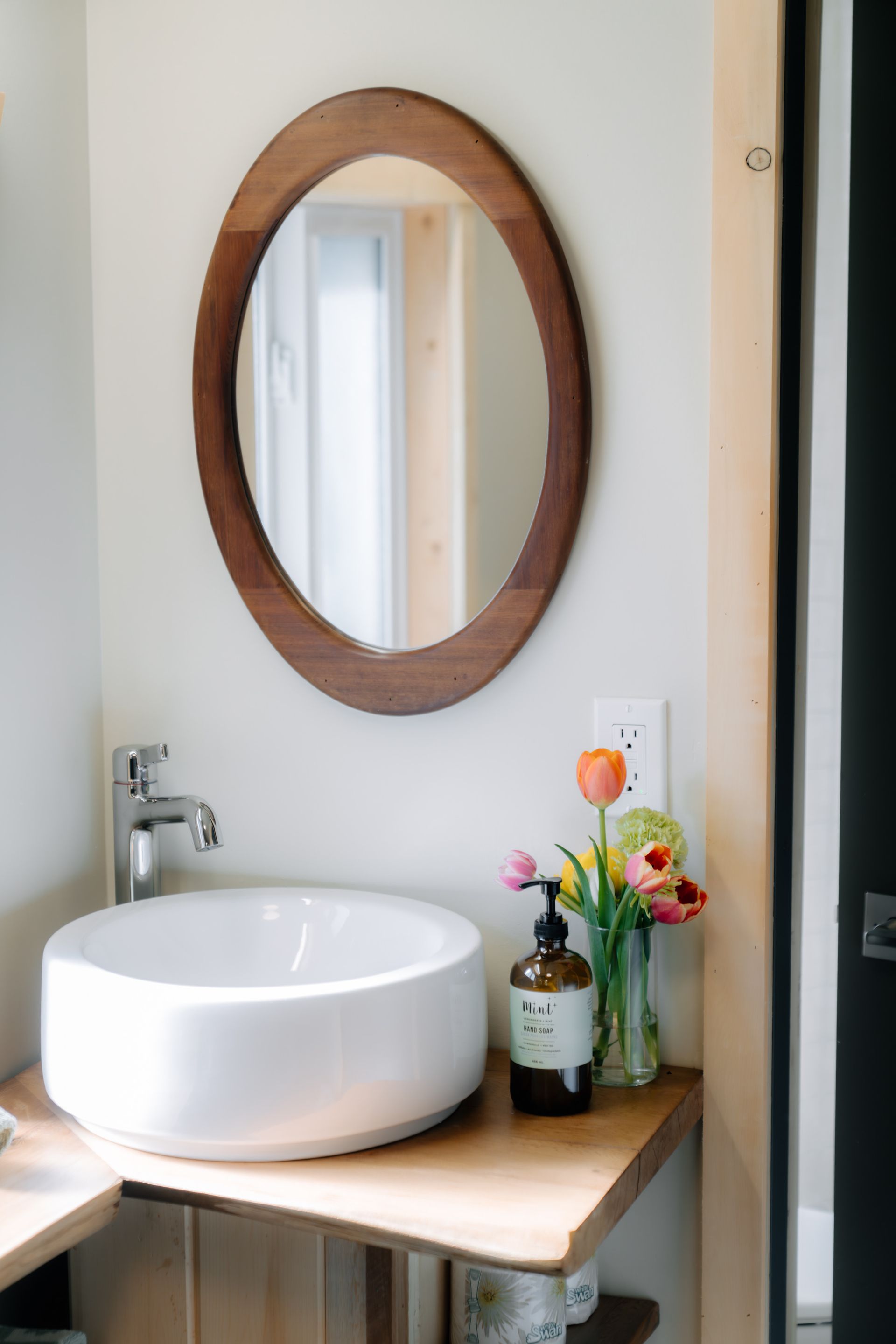 A white vessel sink sits on a wooden counter with a round wooden-framed mirror, soap dispenser, and small vase of flowers.