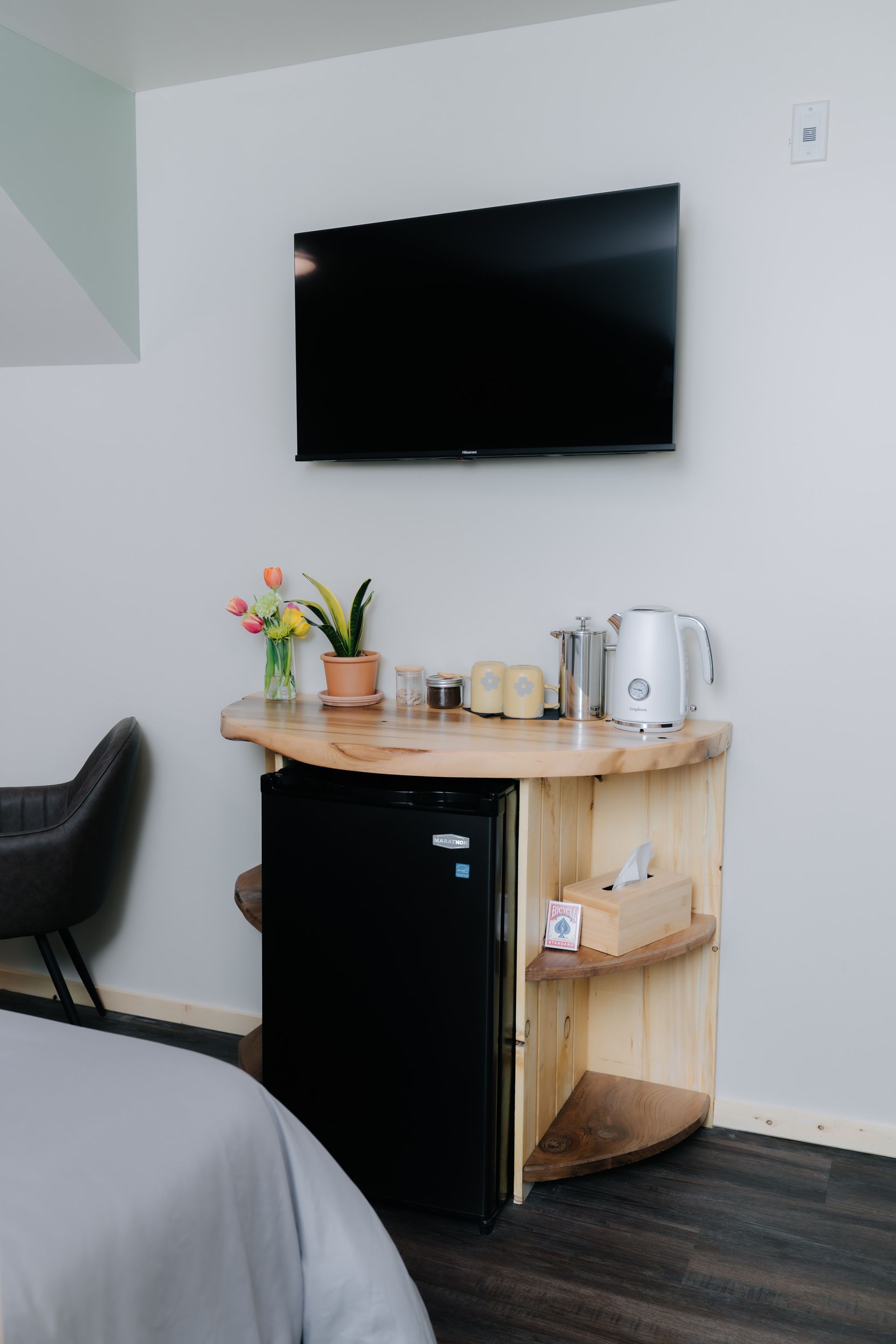 A wall-mounted TV above a custom wooden stand containing a mini-fridge, kettle, and small plant in a hotel room.