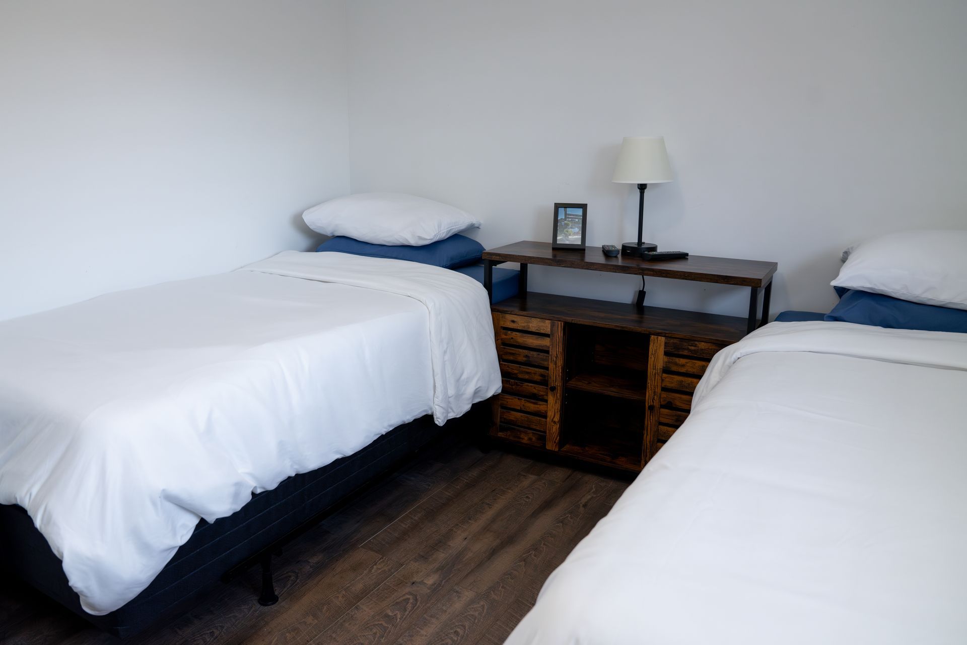 Two twin beds with white bedding and a small wooden nightstand in between against a plain white wall.