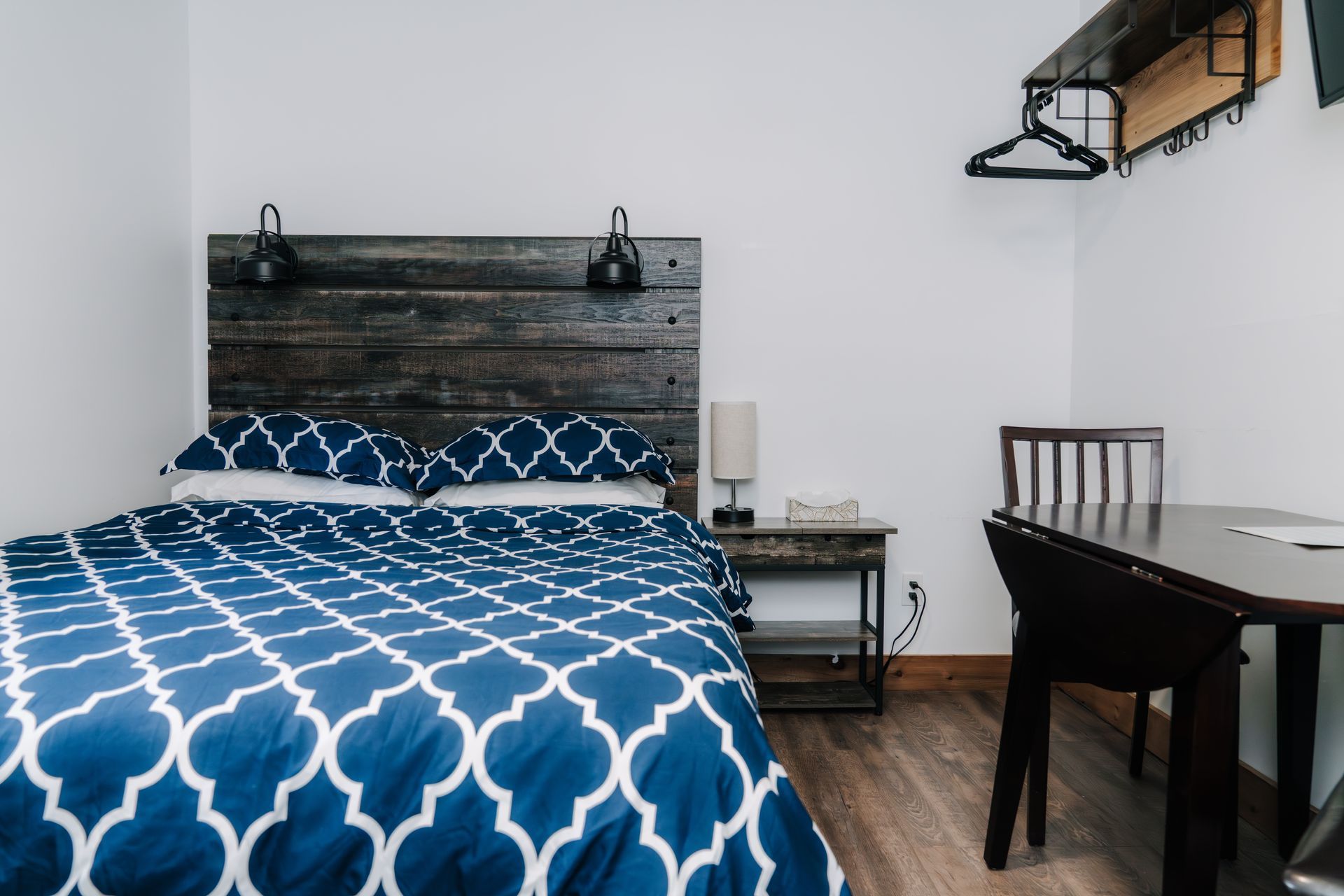 A small bedroom with a bed featuring a blue patterned duvet and dark wood headboard, next to a table and chair.
