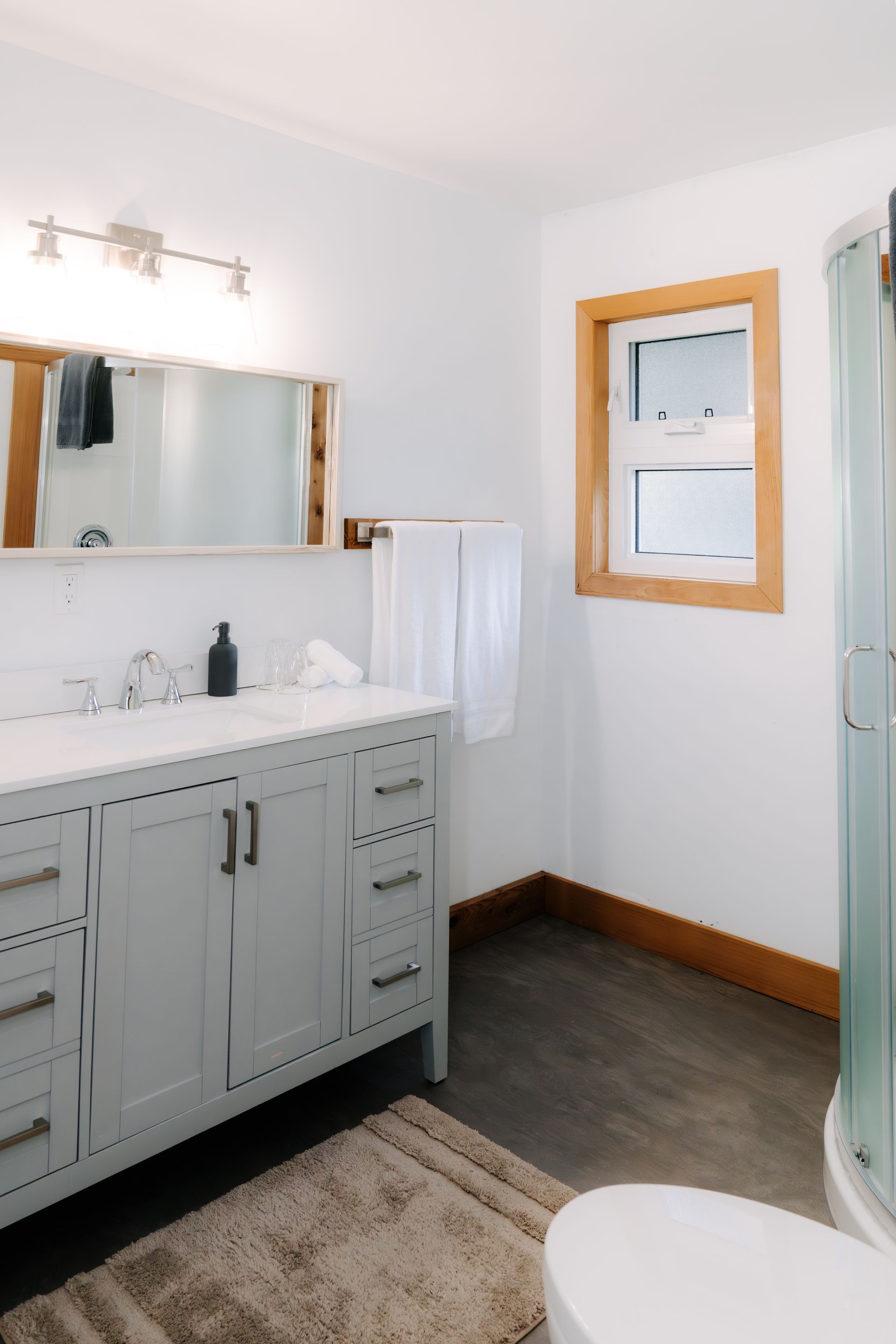 A light grey bathroom vanity with a mirror, white towels, and a window with wood trim, set against white walls.