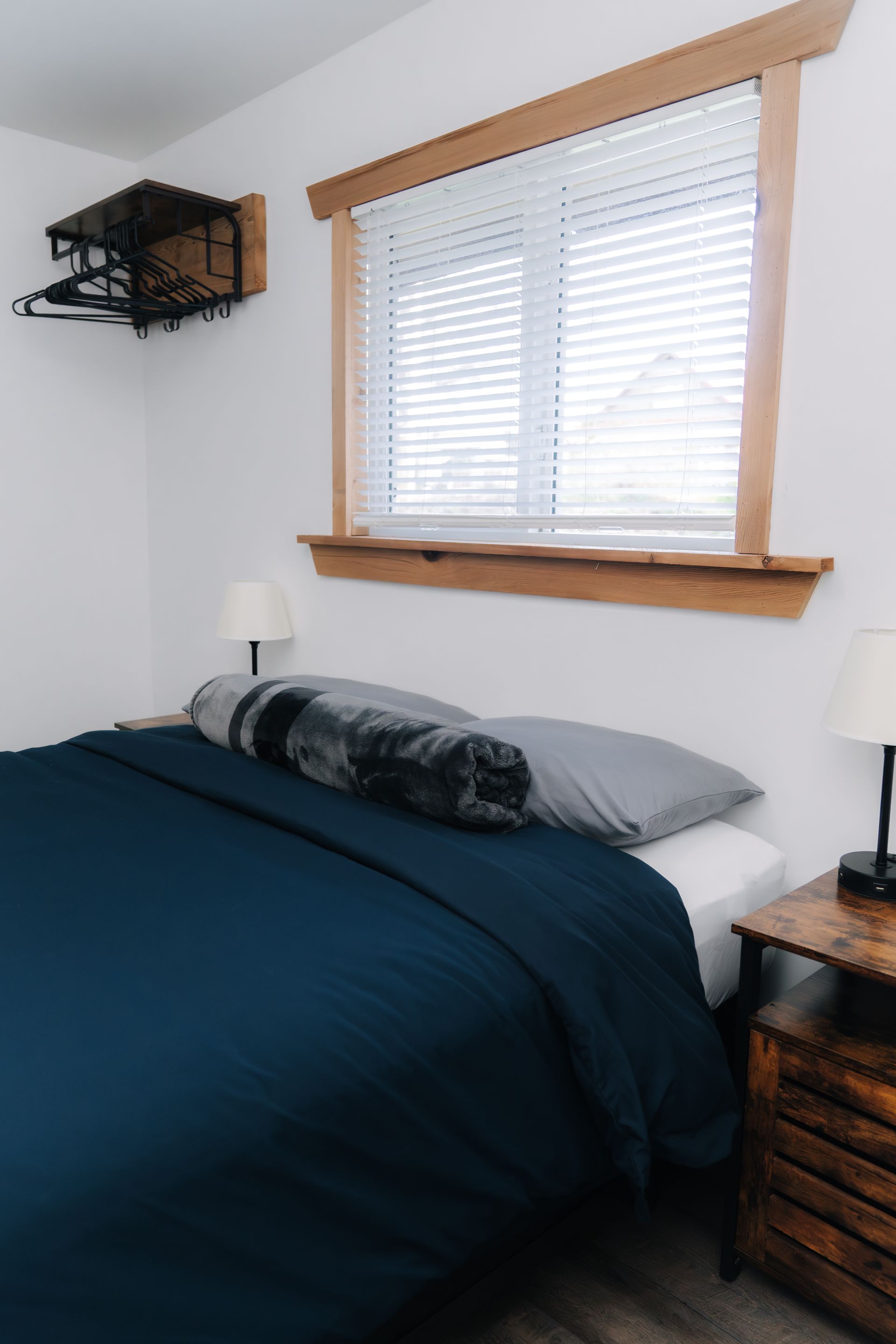 A bedroom featuring a bed with a dark blue comforter, gray pillows, bedside tables, and a wooden window frame.