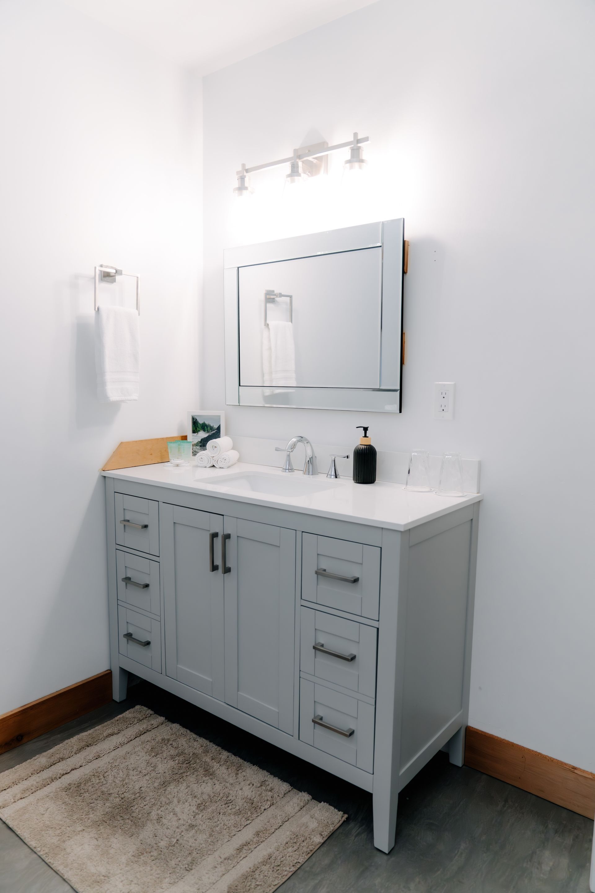 A light gray bathroom vanity with a white countertop, mirror, and wall lighting, set against white walls and wood trim.