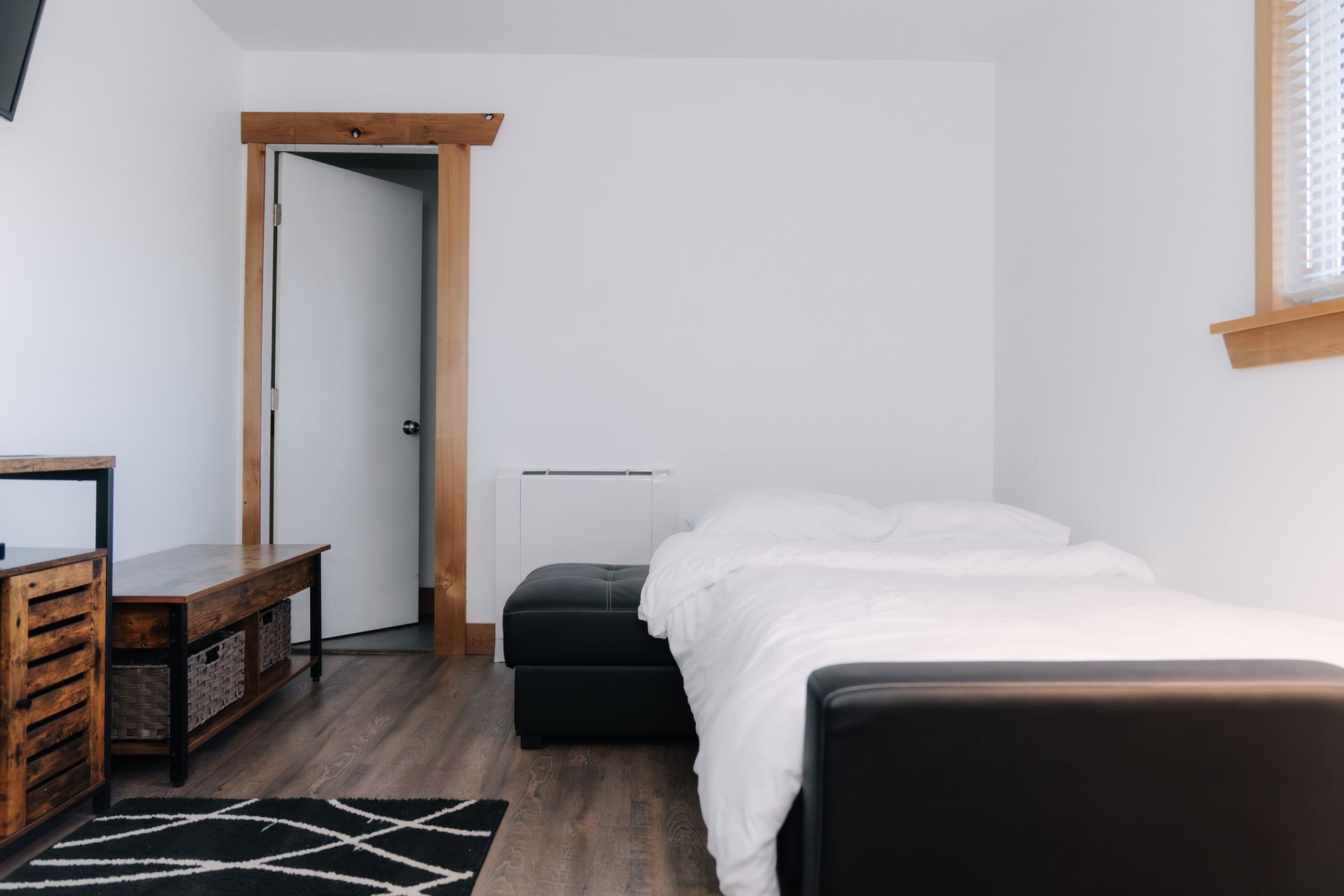 A minimalist bedroom with white walls, a bed with white bedding, a black leather bench, and rustic wood door trim.