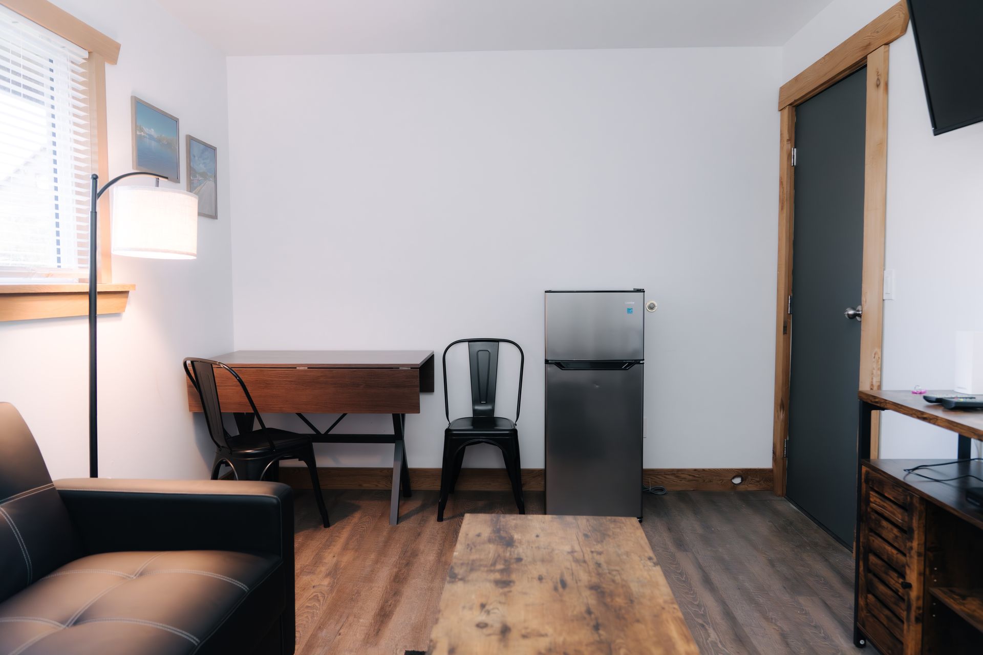 A small hotel room with a leather sofa, a folding desk, chairs, a mini fridge, and a wooden coffee table.