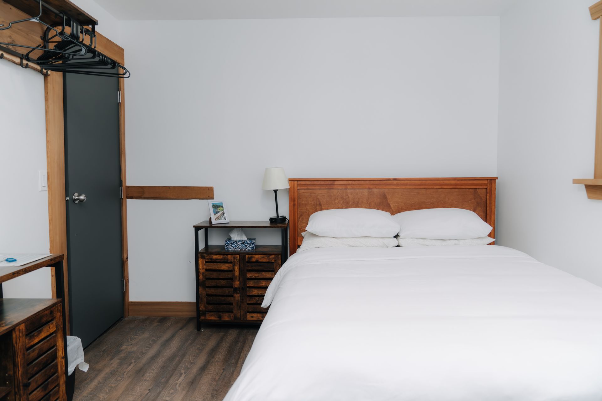 A white-walled bedroom with a wooden bed frame, a white-covered bed, and a wooden nightstand next to a dark grey door.