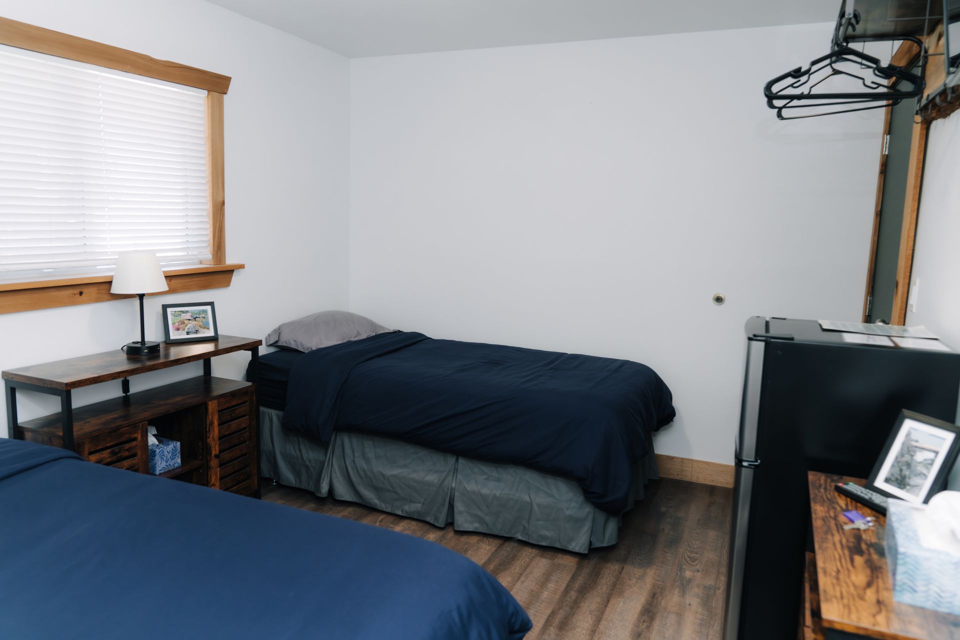 A small bedroom with two twin beds, a wooden dresser, and a small refrigerator against white walls with wood trim.