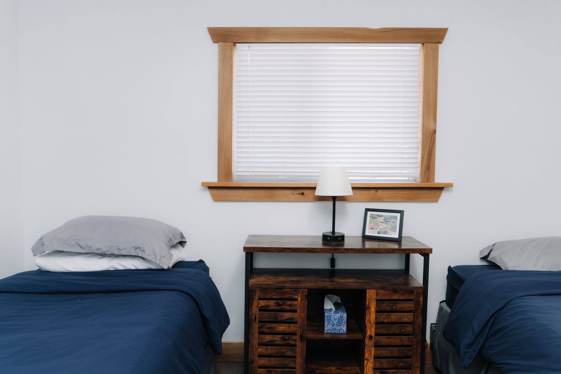 Two twin beds with dark blue bedding and gray pillows flank a wood console table with a lamp and framed photo.