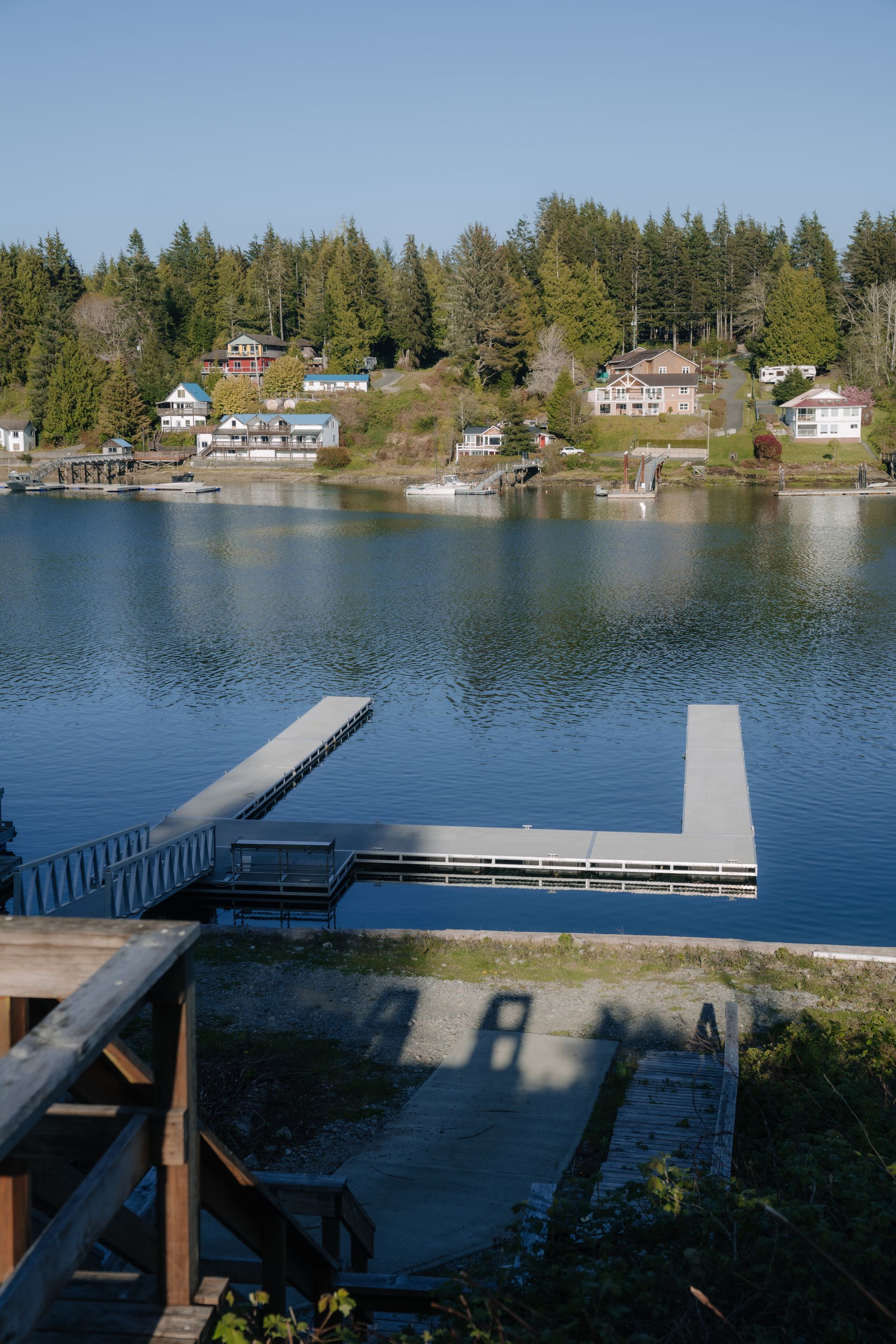 A wooden dock extends into a calm, blue bay with houses and trees lining the distant, hilly shoreline on a sunny day.